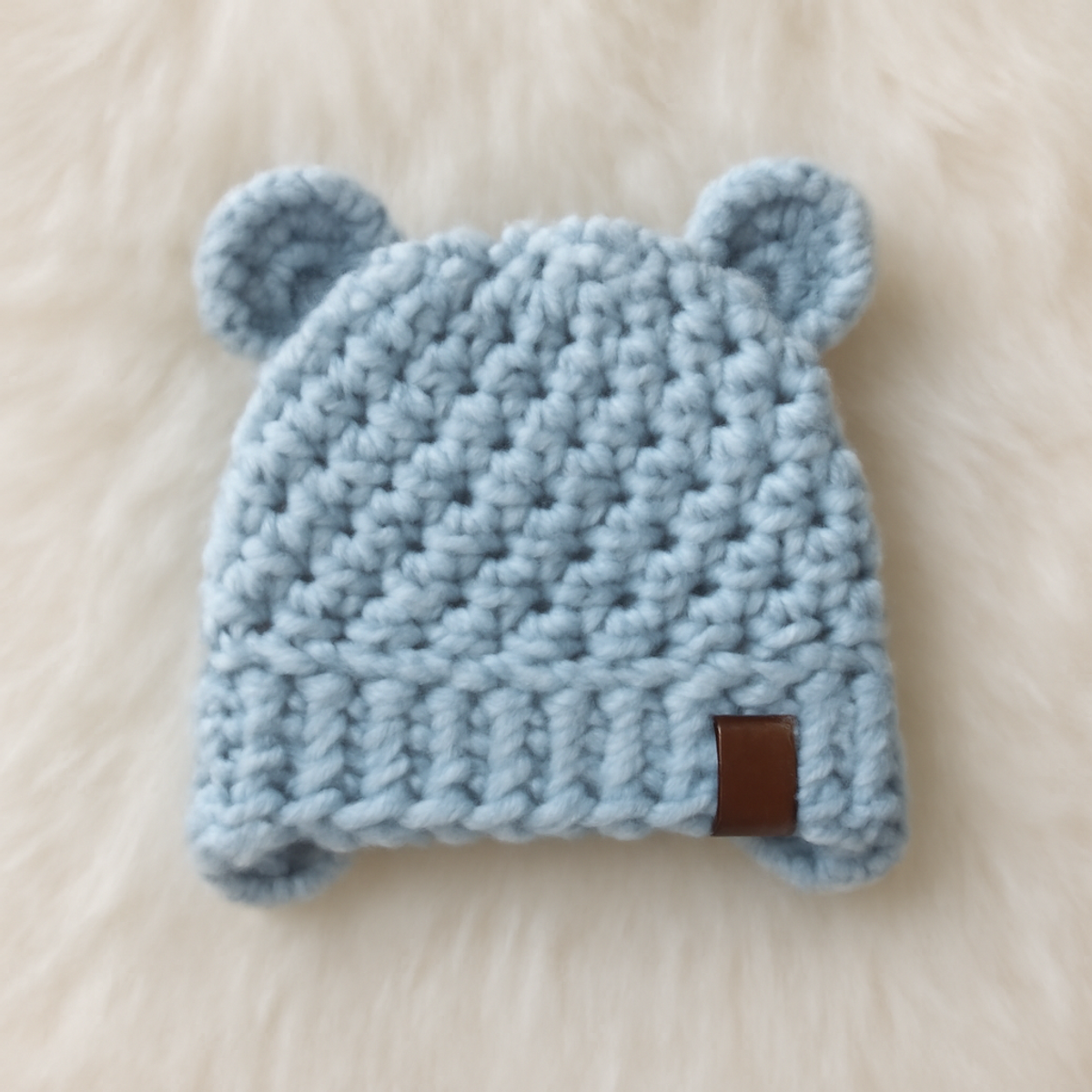 Quick crochet baby hat with bear ears, featuring soft blue and creamy white yarn, warm brown accents, and ear flaps. The hat has a rounded crown and thick ribbing.