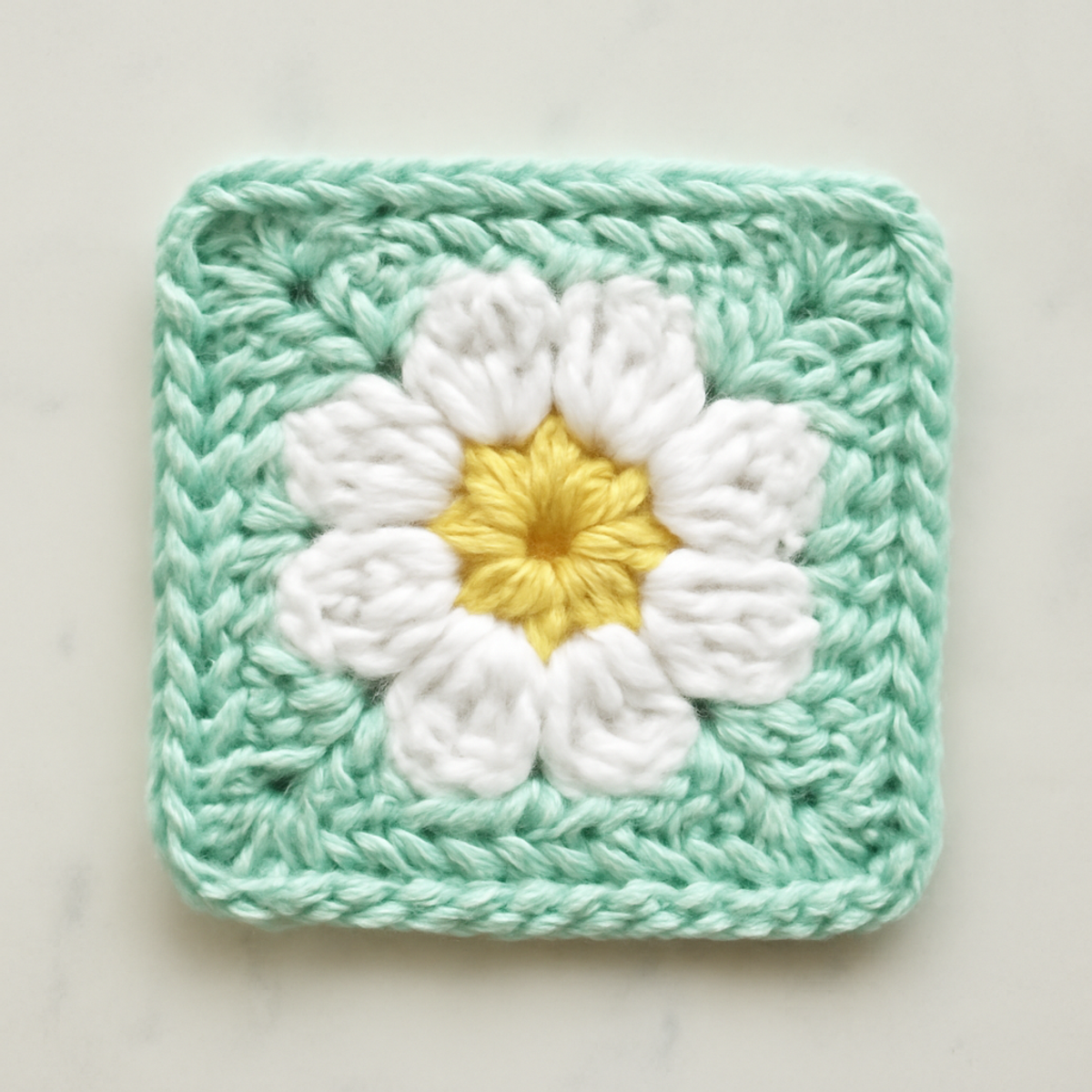 A crochet Dainty Daisy Granny Square displaying mint green, white, and yellow colors in a floral motif with daisy petals and a colorful border.