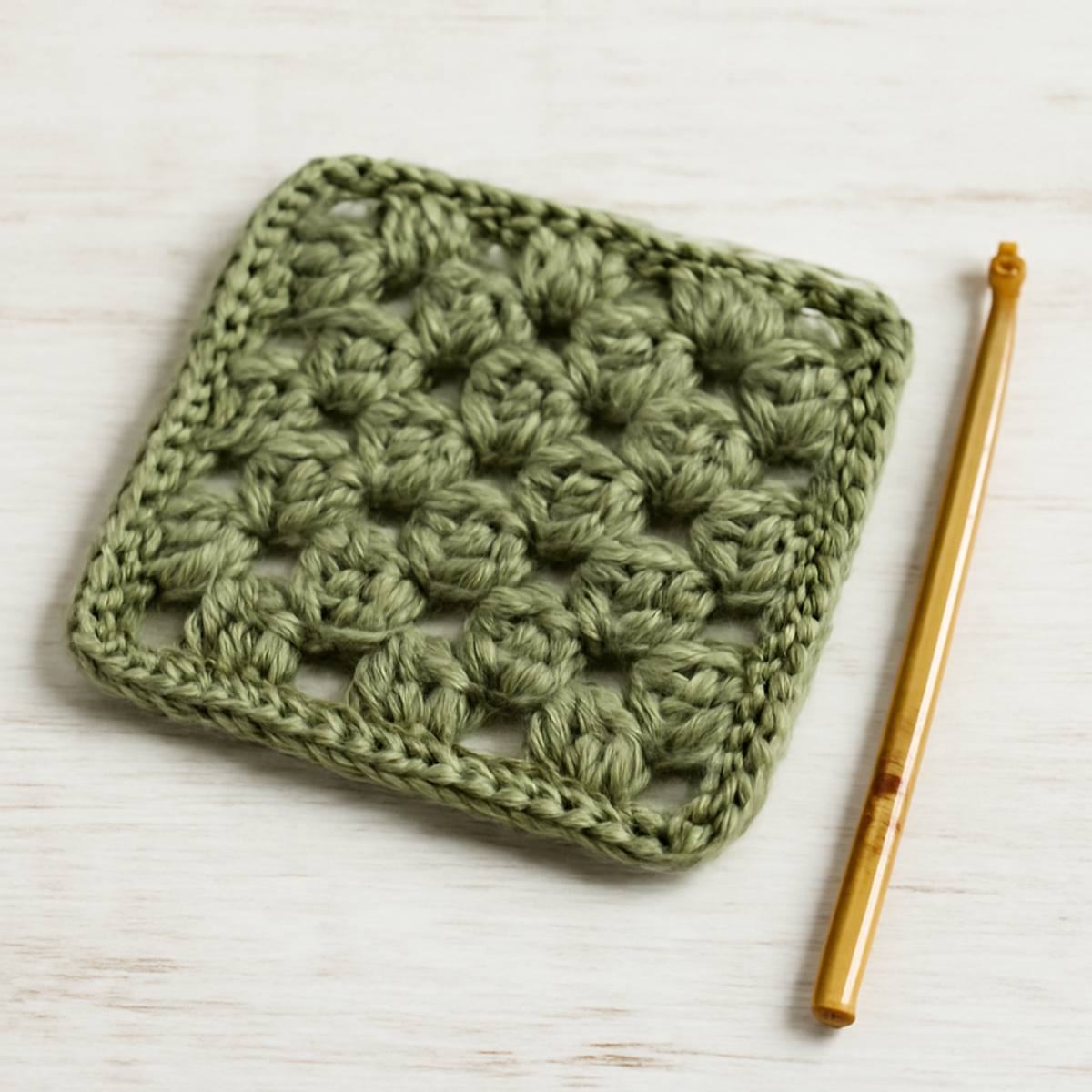 A crochet beginner granny square pattern in olive green showcasing an openwork design with double crochet stitches. The square measures approximately 10 inches by 10 inches.